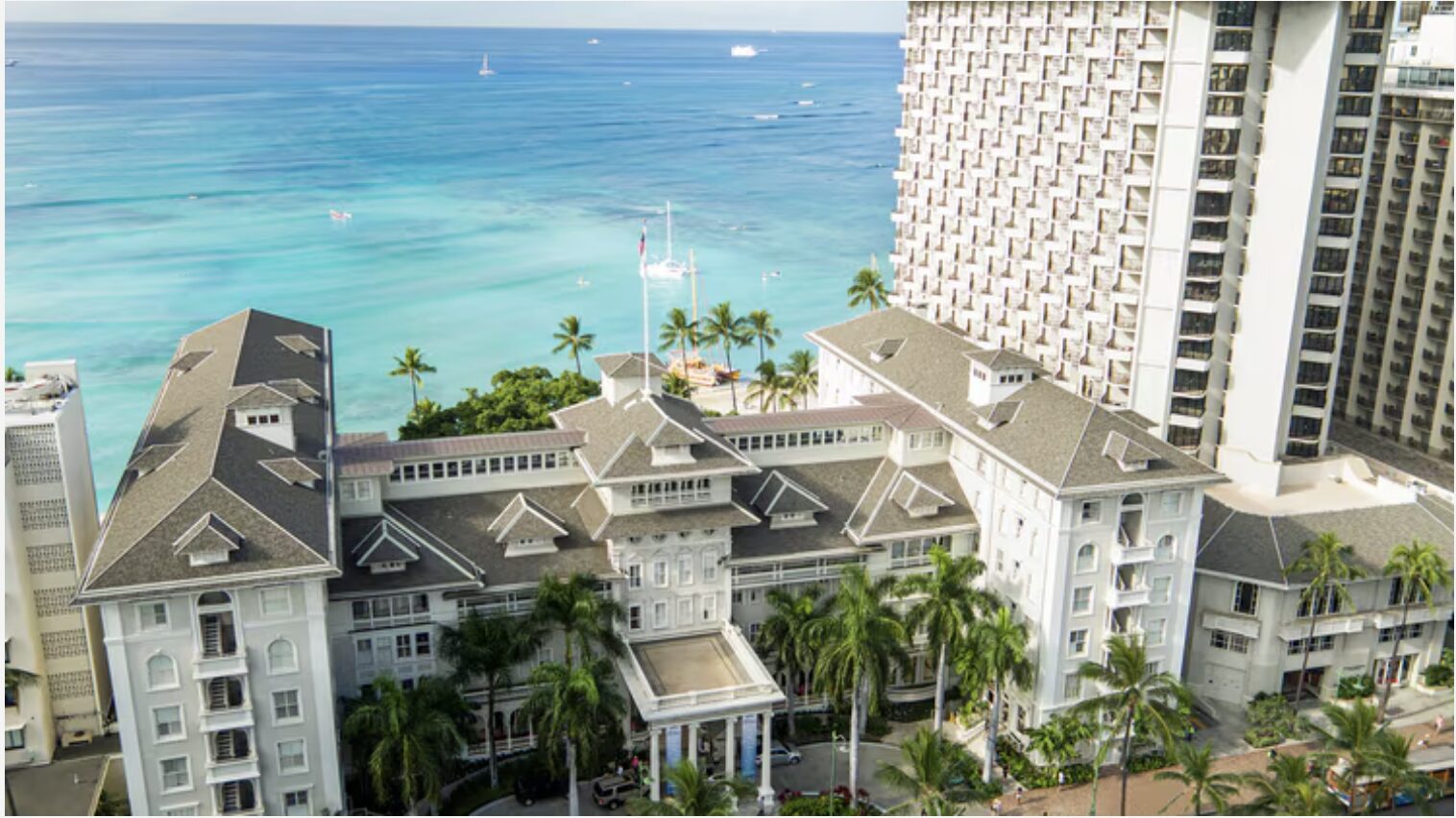 Aerial view of a beachfront hotel with Colonial architecture, featuring palm trees in front. The turquoise ocean stretches into the horizon with a few sailboats scattered across the water. A modern high-rise building is adjacent to the hotel.